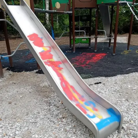 Knowsley Council A slide has been spraypainted with blue, red and yellow graffiti. It is part of climbing equipment which can be seen behind it. Damage can also be seen to safety surfacing behind it, with graffiti spraypainted onto this area too.
