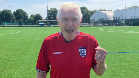 BBC / Jack Hadaway-Weller A person stands on a bright green football pitch wearing a red England jersey. One hand is clenched in a raised fist, and a gold ring is visible on the finger. In the background, there are fenced sports pitches, buildings, and tall light poles under a clear sky.