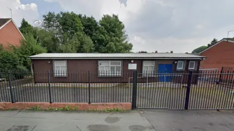 Google A single-storey building with a black fence separating an exercise yard from the pavement. The building has a set of blue double doors and a series of windows and wooden cladding.