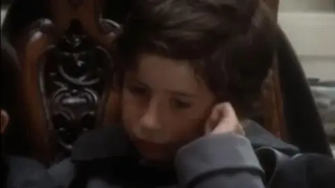 BBC/YouTube A close up shot of John Oliver as a child, dressed in a grey oversized coat and against a wooden ornamental chair