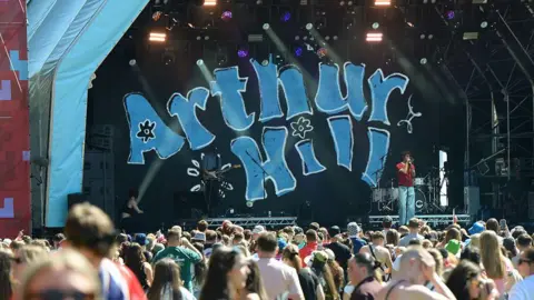 Getty Images A large stage with the name Arthur Hill on the backdrop. The man himself is singing on stage. There is a large crowd milling about in the sun in front of the stage.