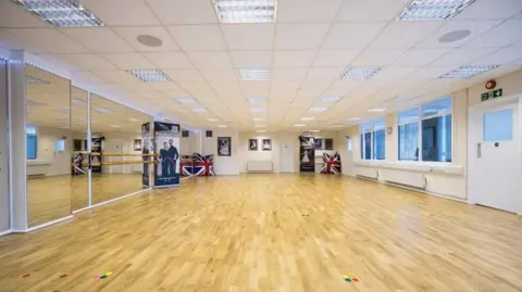 Topline A brightly lit large wooden dancefloor, with windows on one side and floor to ceiling mirrors on the other. There are two large union jack covered cushions in the far right and left corner. 