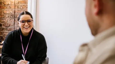 Acquiesce Ltd handout A staff member at the rehabilitation site with brown hair and brown glasses wearing a black top sits in front of a desk during a session with man who has his back to the camera and is blurred out.