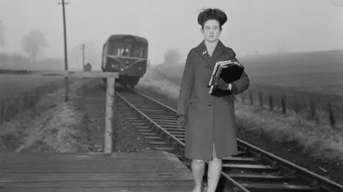 Chris Ware A black-and-white photo of Janet Fisher - then Janet Peare - waiting for the railbus service to Kemble. She is walking up the platform, towards the camera and is carrying some books. She wears a smart coat and has her hair up. A railbus is approaching behind her.