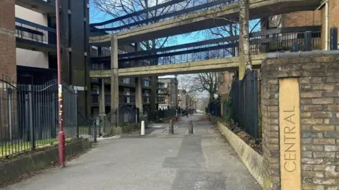 Richard Stead/BBC A walkway under a grey concrete footbridge connecting blocks of flats. The word 'Central' is carved into a stone block in a wall in the foreground. 