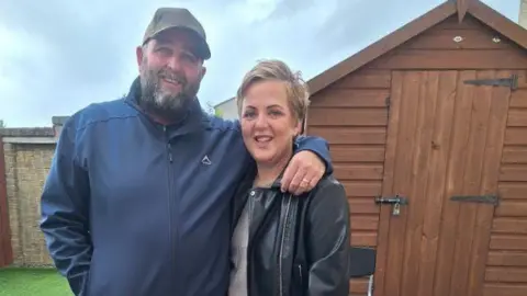 Martin Sutherland A man and a woman are standing in front of a wooden garden shed. The man is wearing a dark-blue jacket and a grey cap, and the blond-haired woman is dressed in a black leather jacket.