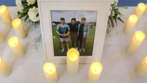 Press Eye A white table covered in a white linen cloth has a photo of one boy, a girl and a woman. There are lit white candles around the fame.