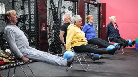 Five members of the class four older and one younger, sit on chairs with their legs outstretched balancing blue balls on their feet