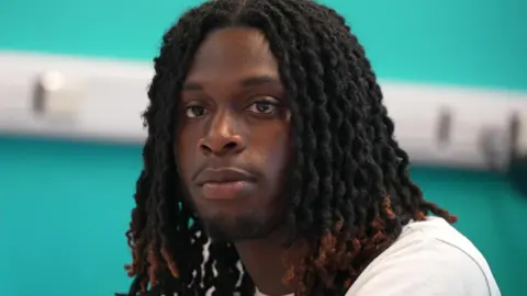 Prince, 18, is wearing a white t-shirt, he is sitting at a table, with a bright blue wall behind him, taking part in the discussion