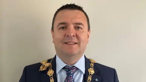 Derry mayor Ruairí McHugh looking directly at the camera and smiling. He has very short, dark hair and is wearing a navy suit, pale blue shirt, navy patterned tie and his gold chain of office.