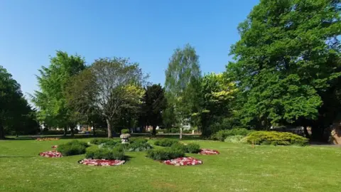 A large open green space in Brenchley Gardens, Maidstone, with mown grass, flower beds arranged in circular displays and a variety of mature trees. Residential buildings can be seen through the trees in the background beneath a clear blue sky.