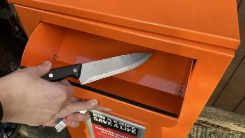 A close-up of a person holding a knife by its handle and placing it inside an orange bin.