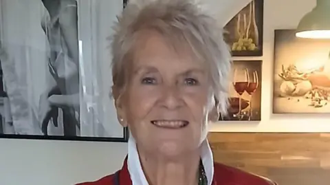 A woman with spiky grey hair and wearing a white upturned shirt is smiling at the camera