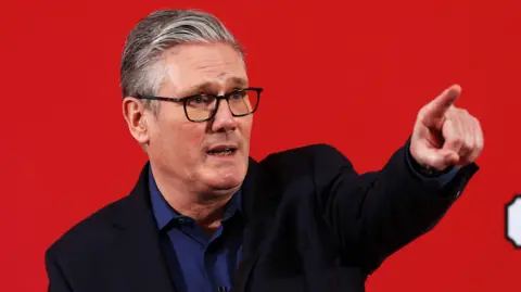 Keir Starmer in a blue shirt and black blazer standing in front of a red background pointing into the distance.