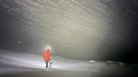 A rescuer wearing a red jacket, black jacket, walking boots and wearing a helmet and rucksack is walking through a blizzard while illuminated by torchlight.