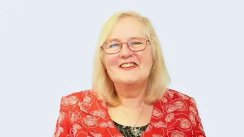 A bespectacled Sue Burke wearing a red patterned top and necklace, She has shoulder length blonde hair and is looking towards the camera, smiling. 