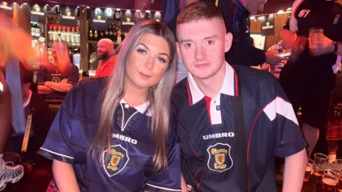 Rachael Vaughan Rachael and partner Aaron wear Scotland football tops and smile together in a pub
