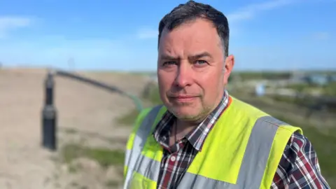 David Balmer, with short dark hair and stubble, wears a checked shift and high visibility vest. He's standing in front of a methane extraction pipe on an active landfill site.