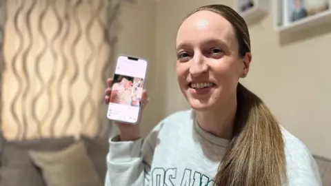 A smiling Stacey Roberts looks directly at the camera as she holds up a mobile phone showing a photograph of her in hospital. She is wearing a grey jumper and has long brown hair tied in a low ponytail. She is standing in a living room with a brown sofa behind her. 