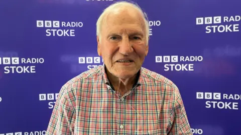 Frank Blunstone, an elderly man with short white hair and a red chequered shirt, smiles as he stands in front of a purple BBC Radio Stoke-branded background.