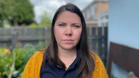 A photo of Kirsteen Buckney looking directly at the camera. She is outside in a back yard. She has long dark hair and wears a yellow cardigan over a blue shirt