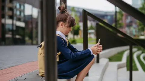 A young girl in a blue dress is sitting on steps outside a school building looking at a smartphone. 