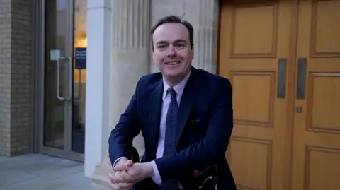 Gregory Burke A man smiles at the camera, with his hands clasped in front of him, and wears a navy suit, pink shirt and blue tie with small pale dots. Behind him is a stone building, with wooden doors directly behind him.
