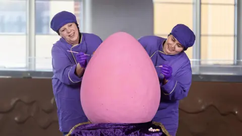PA Media Two women in purple overalls and head coverings stand on either side of a giant pink mini egg, holding paint brushes. Behind them a fake melted chocolate wall can be seen, and windows.