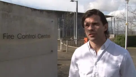 Riccardo la Torre, who has long brown hair and is wearing a white shirt. He is standing in front of a building outside on a cloudy day. To the left is a sign that reads "Fire Control Centre".