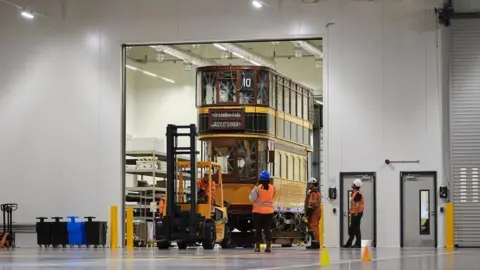 Science Museum Group Inside a warehouse-type building, a large doorway with a forklift pulling through a yellow and brown vintage tramcar with two levels