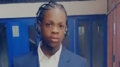 Tyler McDermott, a young man wearing a blue jacket and white shirt while stood in front of a blue wall and doors