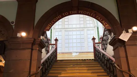 An image of the main staircase - an impressive wooden construction with fancy ironwork. The words "Welcome to the People's Palace" arch over the stairs in a light gold.