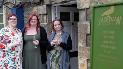 Three women standing outside a stone doorway beneath a green ‘Jackdaw Gallery’ sign, each holding a drink; the gallery entrance and signage are visible behind them.