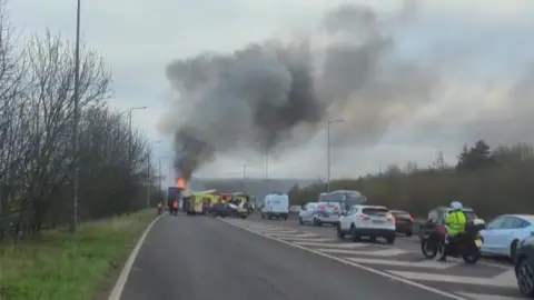 A fire crew uses a high-powered hose to dampen down a smoking lorry, which was on fire. There is a fire engine in the foreground.