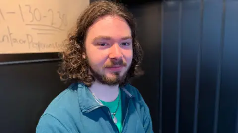 A young man with long hair and a beard wearing a green-blue fleece and a green t-shirt