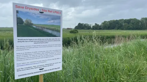 A sign says "Save Drysides" in red writing with a description of the area below. In the background is the old River Nene, with long grass either side and a field and copse in the backdrop.