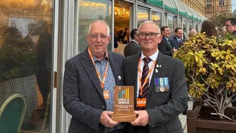 Richard Thomson Two men stand outside the House of Commons in London, holding a wooden plaque that reads “Men’s Sheds Armed Forces Communities Award 2025”. One wears military medals, both have visitor badges.