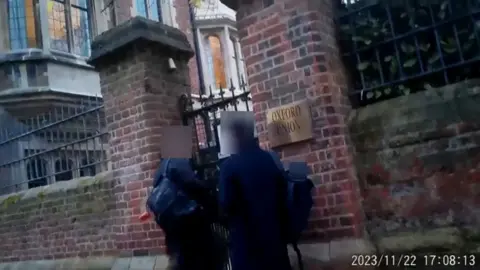 Counter Terrorism Policing Video footage of two men with blurred faces, dressed in black and carrying rucksacks, standing outside the entrance to the Oxford Union. 