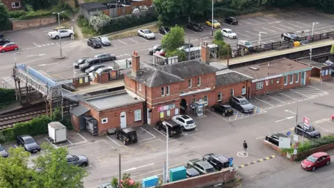 An aerial view of a railway station and surrounding car parks. A red‑brick station building sits near train tracks, with platforms on either side connected by a covered footbridge.