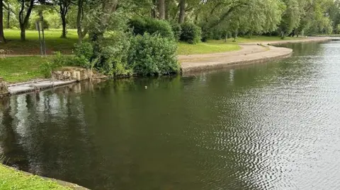 Blackpool Council Looking across Stanley Park lake which is a dark green colour with trees and a winding path to the left and a small fishing platform