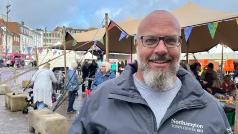 Oliver Conopo/BBC Man with grey beard, glasses and bald head smiling at camera, wearing a light grey tshirt and darker grey zip-up jacket with "Northampton Town Centre Bid" in white lettering to one side of the chest. Behind him and obscured by him is a stall covered with an awning and surrounded with coloured bunting and various people standing around and buildings around the market square.