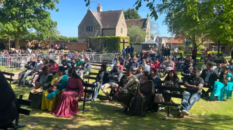 Richard Knights/BBC Lots of people are sat on benches in rows on a green field outside of the chapel. In the background is a stone house and two trees.