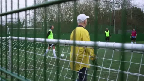 Man in yellow shirt guards goal whilst players run with ball in the background. The game goes on behind a mesh fence.