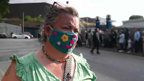 Aisha Iqbal/BBC A woman wearing a colourful face mask and a green cotton top and beaded necklace. Behind her out of focus stands a crowd of people.