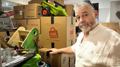 BBC A man look back at the camera as he gestures with one hand towards a pile of boxes, a child's green dinosaur chair, a seed spreader and plastic bags for life. He has short white hair and a clipped salt and pepper beard. he is wearing a linen elbow length shirt open at the neck 
