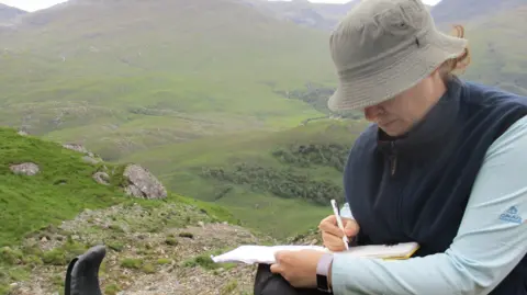 Claire Dashwood Claire Dashwood wearing a brown bucket style hat, navy gilet and baby blue long-sleeved top, writing in a notepad. She is sitting on a cliff and there are rolling green and rocky hills behind her. 
