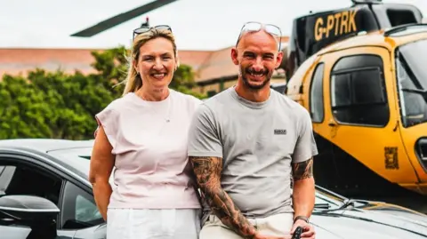 Elite Competitions A man and woman are seen sat on a car. The woman has a pale pink top and blonde hair with sunglasses on her head. The man has a grey top, tattoos on his arms and sunglasses on his head. They are both sat in front of a yellow helicopter