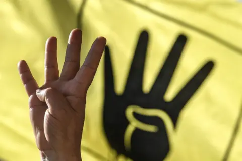 AFP A man's hand is seen gesturing with four fingers held up. A black-and-yellow banner behind him depicts a hand making an identical gesture.