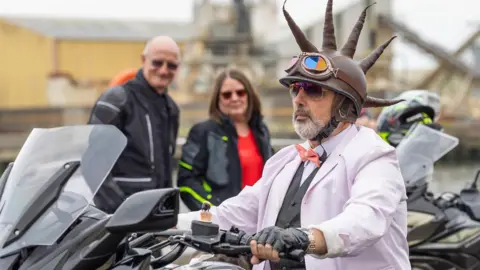 A man in a lilac suit, bow tie and a helmet with punk spikes riding a motorbike.
