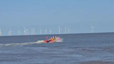 Supplied An inshore lifeboat - a small, orange craft carrying three people wearing yellow jackets, red lifejackets and white helmets - makes waves as it heads out into a calm-looking blue sea under a blue sky, with white wind turbines on the horizon.